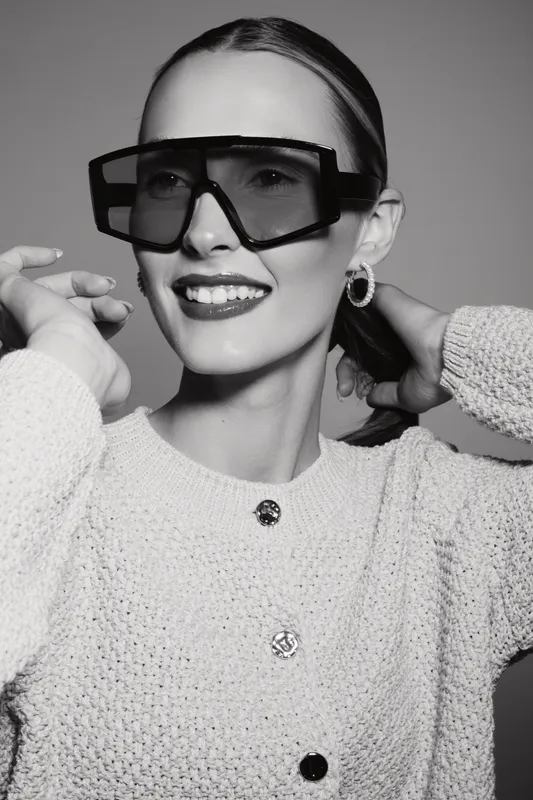 Smiling white woman with light skin and slicked hair wears large sunglasses and textured sweater, posing confidently in studio. Concept of fashion, style, and modern elegance.