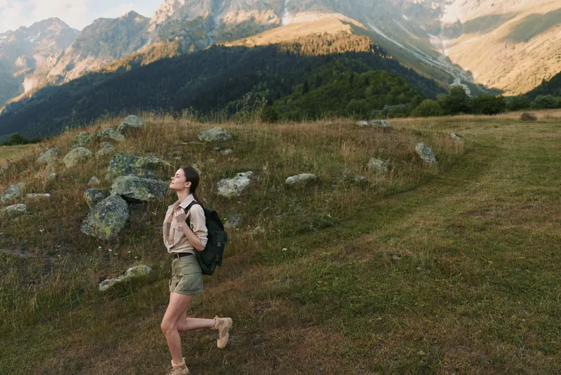 Young woman hiking along a mountain trail with a backpack across an open meadow, capturing adventure, exploration, and a calm, confident mood, with natural light highlighting the rugged landscape