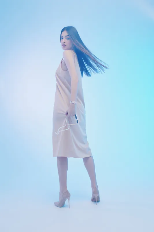 Young woman in a beige dress and high heels poses with sunglasses in hand on a soft blue background. Concept of beauty, fashion, elegance, and style.