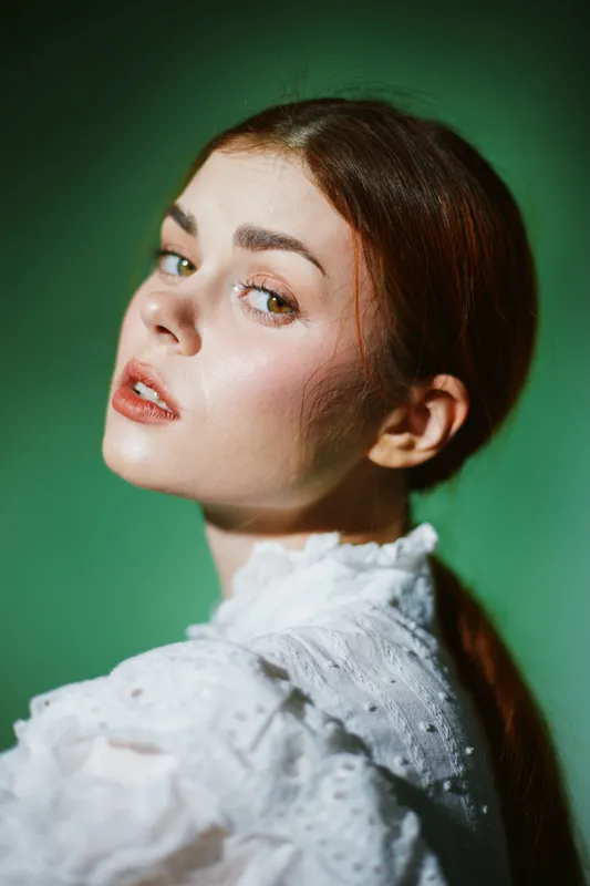Young woman in vintage white blouse with soft expression against a green background, showcasing natural beauty and elegance in a seasonal portrait