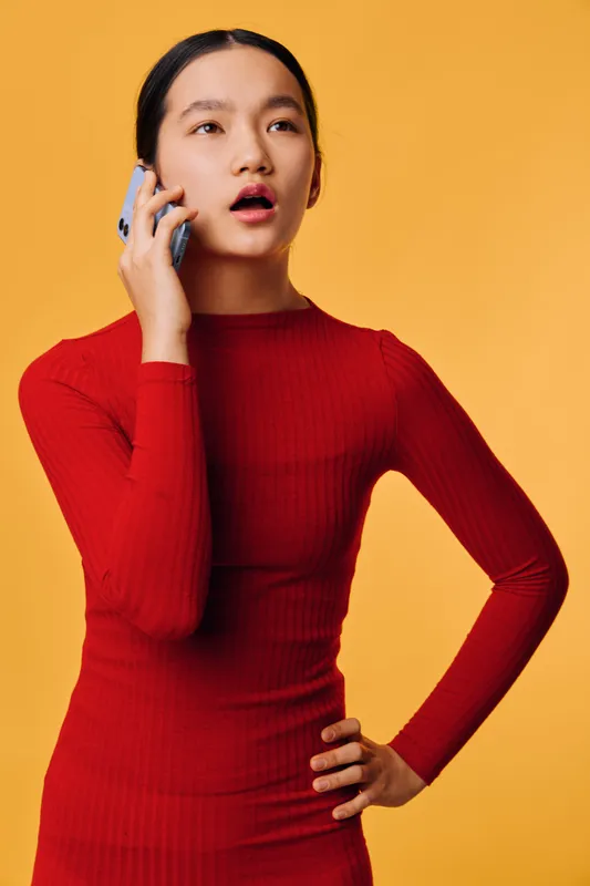 Young woman talking on smartphone with worried expression standing against yellow background wearing red long sleeve top. Communication, emotion, technology and lifestyle concept.
