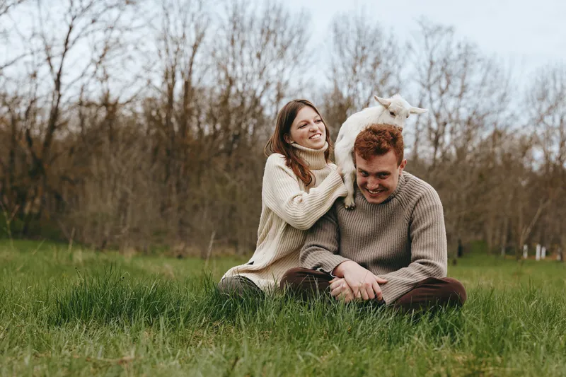 Young couple wearing knitted sweaters sits on grass in a field while a playful baby goat climbs on the man's shoulders during a relaxed lifestyle moment.