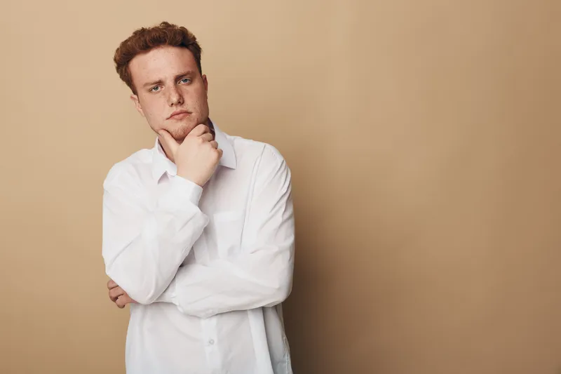 Thoughtful young man in white shirt stands against beige background with hand on chin. Concept of contemplation, decision making, and casual style for lifestyle and portrait.