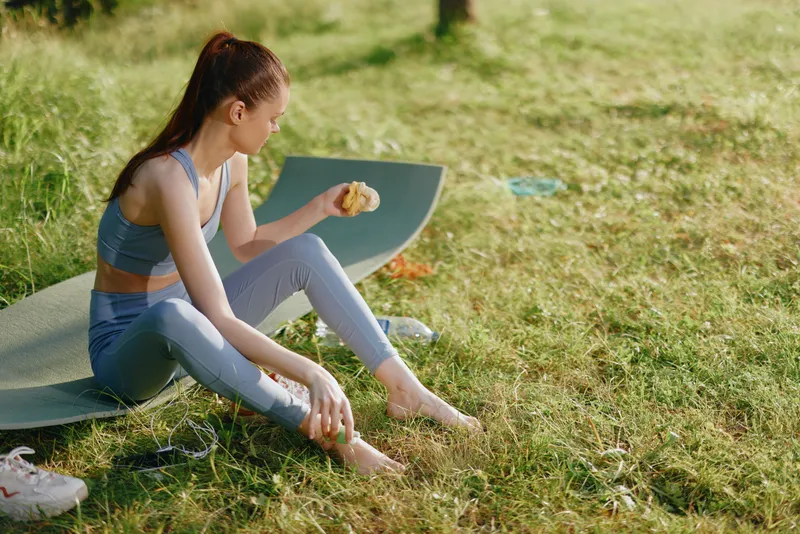 Young woman enjoying a healthy snack on a yoga mat outdoors during a sunny day, promoting healthy lifestyle and fitness