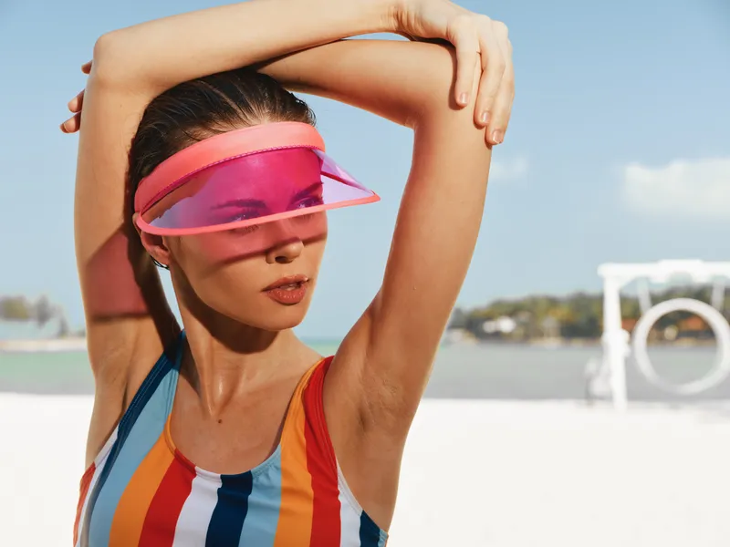 A stylish woman on a sunny beach wears a colorful striped swimsuit and a pink visor, striking a relaxed pose with arms raised, capturing a vibrant summer vibe.