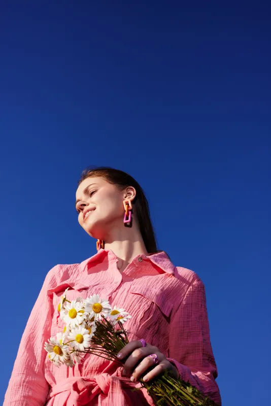 Young woman in a pink outfit holding daisies against a clear blue sky, radiating joy and positivity during springtime