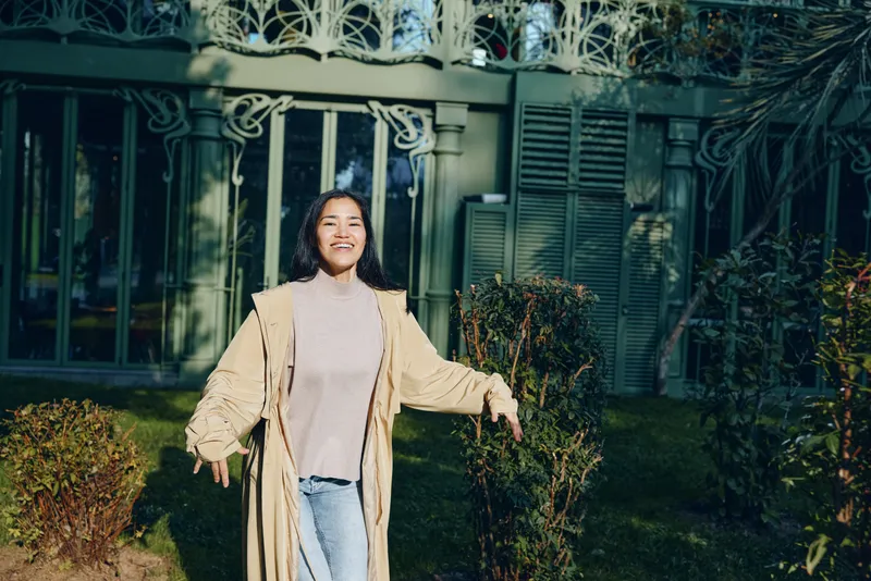 Woman standing in a garden, open coat, smiling under sunlight, casual fashion, outdoor scene with greenery, relaxed pose, natural light, cheerful mood, everyday style, candid moment in a serene yard