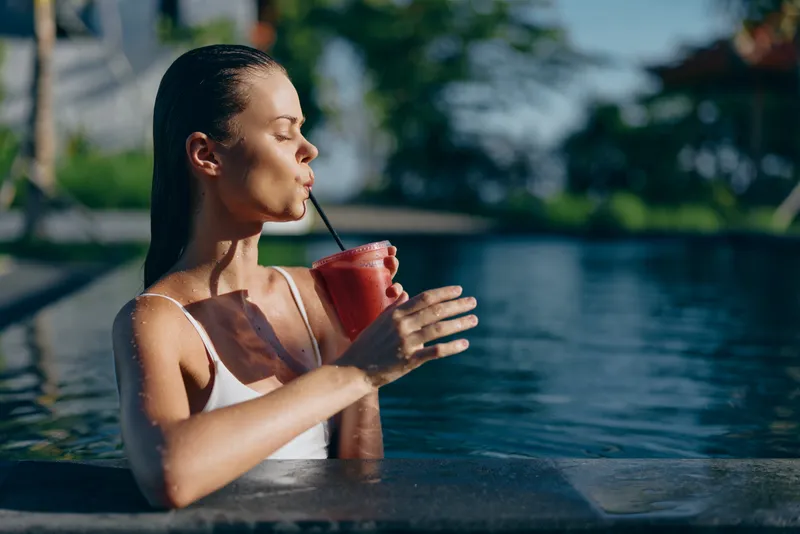 Woman relaxing in a pool drinks a pink smoothie, enjoying sunny moments poolside, casual swimwear, calm mood, clear water, lush background, vibrant colors, lifestyle photo