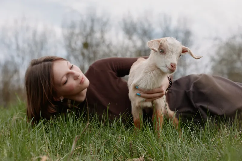 Lifestyle scene with a woman lying on lush grass, a goat kid nearby, soft light, candid expression, natural colors, peaceful outdoors, gentle affection and connection in a rural meadow