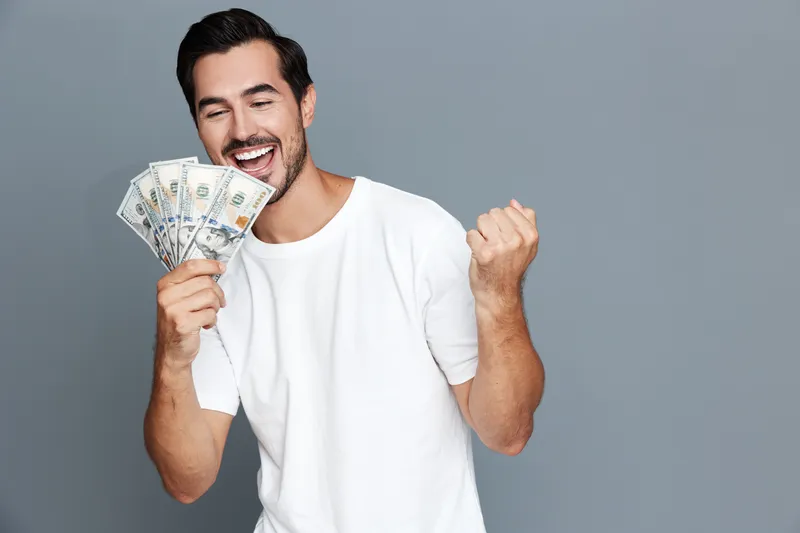 Happy cheerful young man holding money in hand, celebrating success with excitement isolated on solid gray background. Emotion and finance concept.