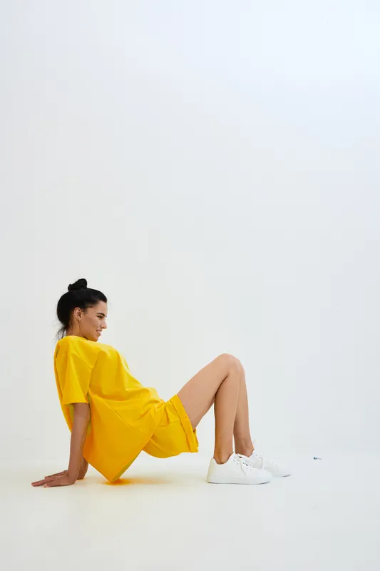 Young woman in yellow dress sitting on floor against white background, casual style, relaxed pose, modern fashion, clean minimalistic interior setting.