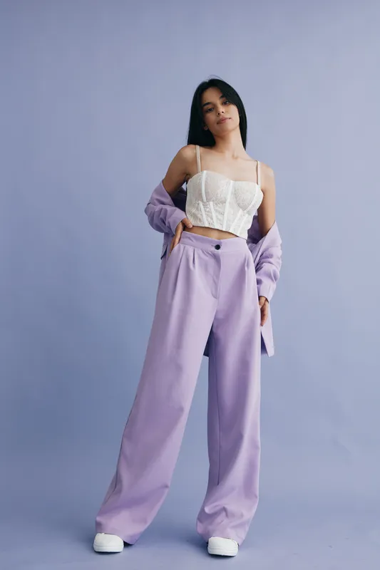 young woman wearing lace white top and lilac wide-leg pants poses confidently against lilac background, showcasing stylish casual fashion in soft pastel tones