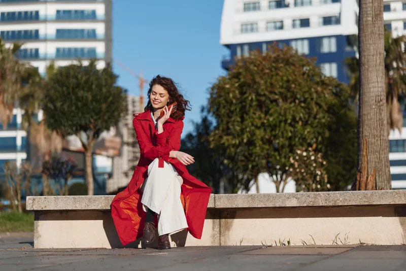 Young woman in a red coat sits on a bench outdoors in a city park enjoying the sunny autumn day with a relaxed and content expression.