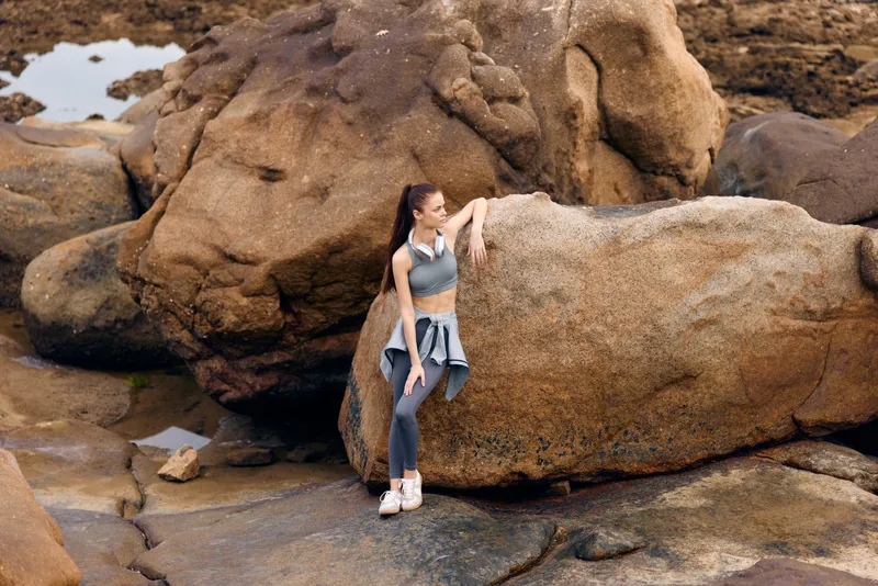 Young athletic woman in workout attire poses confidently on rocky coastline, showcasing strength and natural beauty in outdoor fitness setting