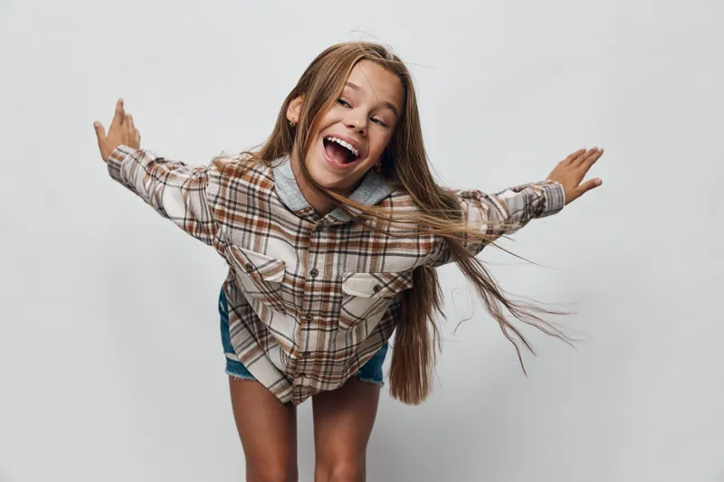 Girl in plaid shirt with long hair smiling and spreading arms wide on white background. Happy teenager, casual style with joyful expression and playful pose for youth energy.