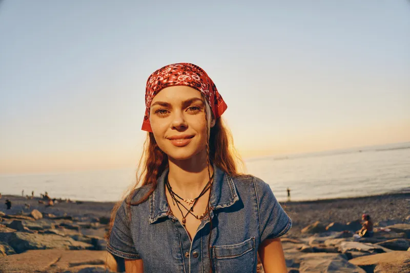 young woman with red bandana smiles warmly on rocky beach during golden hour sunlight, casual denim shirt and layered necklaces create natural, relaxed vibe