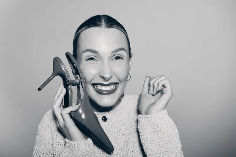 Young white woman with shiny hair smiles holding high heel shoe near face in cozy sweater studio.