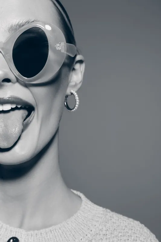 Young white woman with pale skin wearing large round sunglasses sticks out her tongue. Close-up portrait in black and white with playful and rebellious mood on grey background.