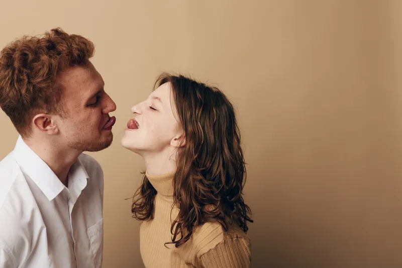 Young couple with curly hair playfully interacts, woman sticking out tongue, man leaning in close. Casual clothing and neutral background create intimate, fun, and lighthearted mood.