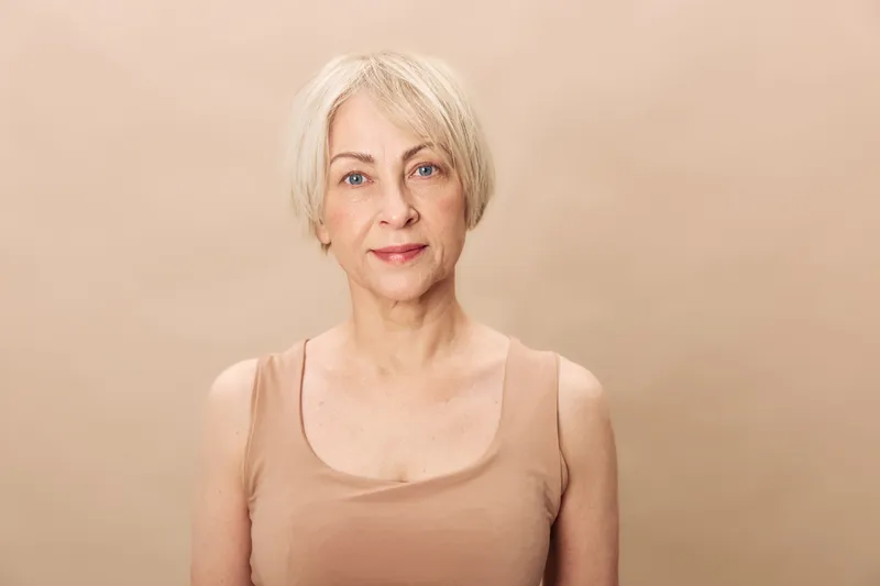 Portrait of a mature woman with short blonde hair and blue eyes wearing a beige tank top. Neutral background emphasizes natural beauty and aging gracefully concept with soft lighting.