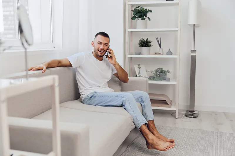 Smiling man enjoying a phone conversation while relaxing at home in a modern, airy living space. Comfortable atmosphere with light colors and greenery.