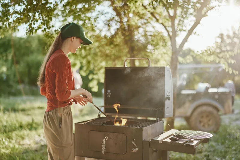 Young woman grilling outdoors in a summer landscape, wearing casual clothes and a cap, surrounded by greenery, showcasing joy and relaxation while cooking