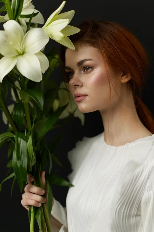 Young woman with red hair holding white lilies, exuding calmness and beauty with a dark background, perfect for seasonal and wellness themes