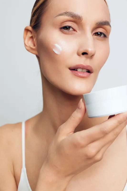 Young woman applying white cream on her cheek holds jar skincare product close to face with natural fresh skin and neutral expression isolated on light background