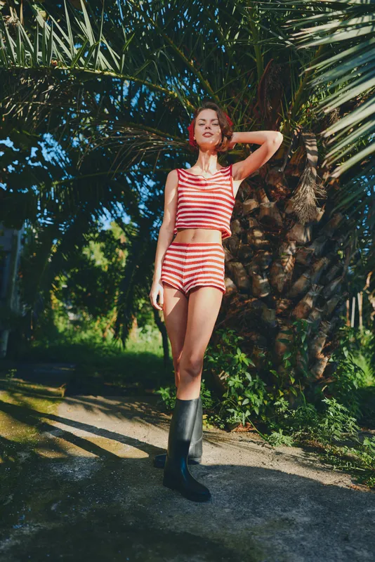 A confident woman wearing a red and white striped swimsuit stands beside a palm tree in a sunlit outdoor setting. She strikes a relaxed pose, exuding summer style and confidence.