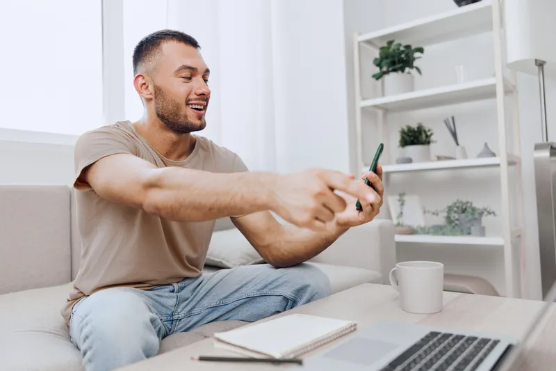 Happy man using smartphone while relaxing at home, enjoying communication and the comforts of modern technology in a cozy living room setting.