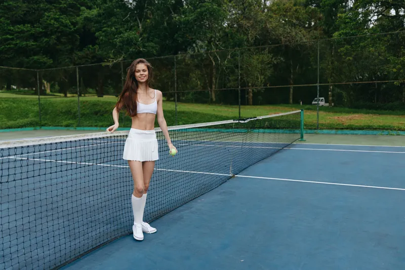 Young female tennis player in sporty white outfit, smiling on the tennis court, showcasing athleticism and joy in a lush green environment