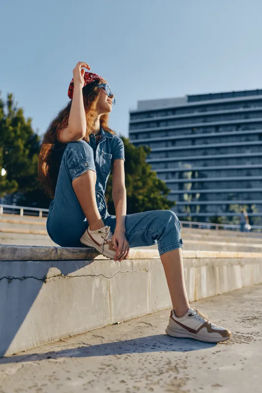 Lifestyle woman in modern boho-western denim street style sitting outdoors in casual sneakers under soft natural sunlight with film color effect enhancing warm tones.