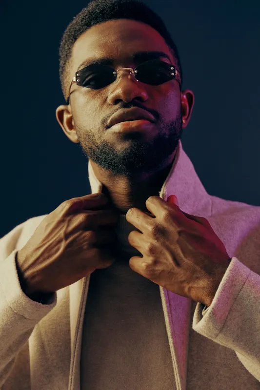 Stylish young man in sunglasses poses confidently against a dark blue background, showcasing modern fashion and sophistication with a trendy outfit