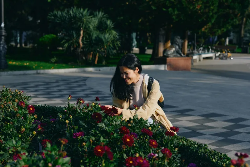 Woman in a yellow dress tends vibrant flowers in a city park, sunlight filtering through trees, a serene outdoor gardening moment, colorful fragrance, and floral beauty