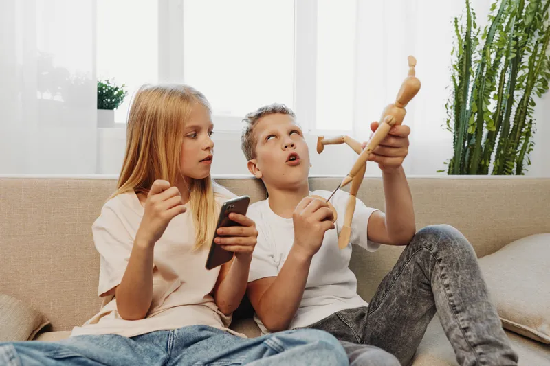 Two children sitting on a sofa play with a wooden mannequin and a smartphone in a bright living room with natural light and plants in the background.