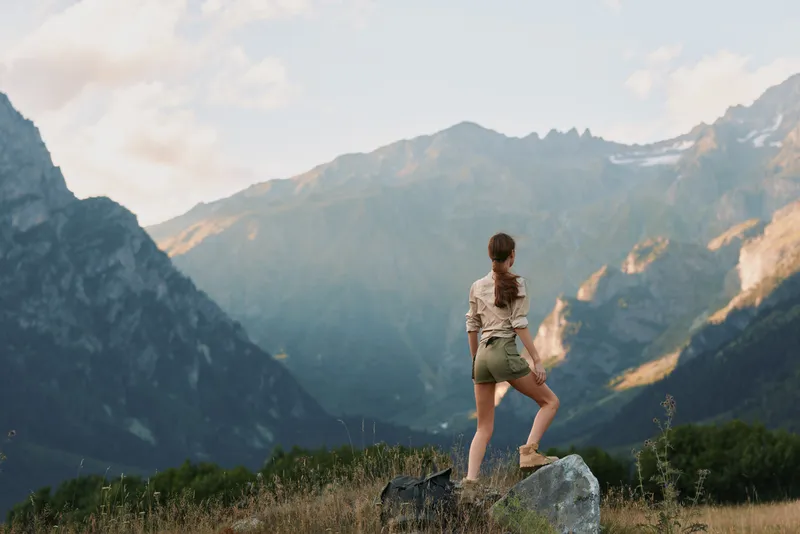 Young woman hiking in mountains, standing on rock, enjoying scenic view, casual outfit, outdoor lifestyle, natural light, summer day, adventure spirit