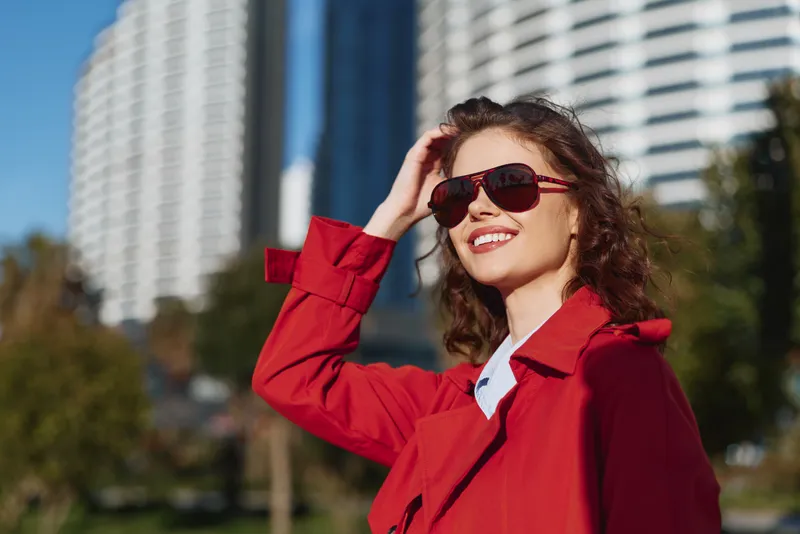 Happy woman wearing red coat and sunglasses outdoors, smiling brightly, sunny day, urban background, casual style, confident expression, natural light
