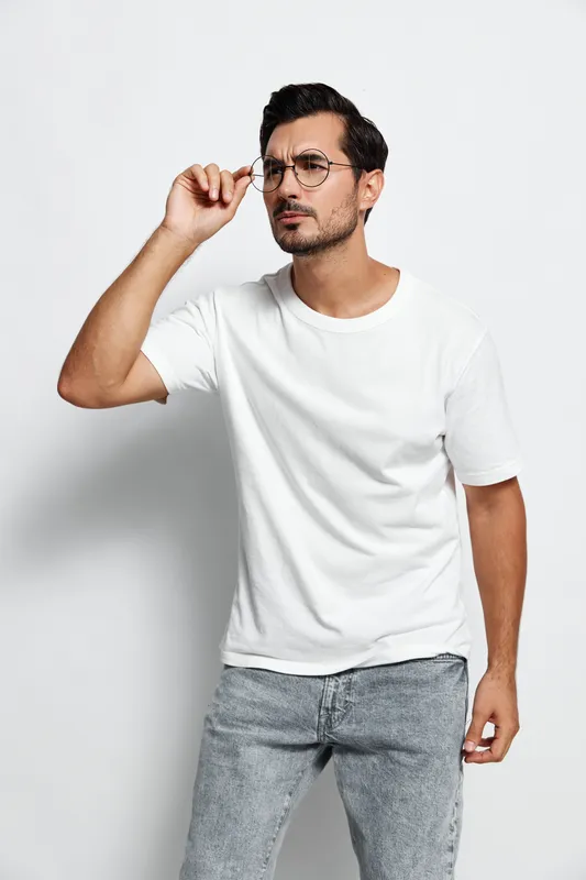 Thoughtful man adjusting glasses while looking into the distance. He wears a casual white t-shirt and blue jeans against a plain light background. Personal expression concept.