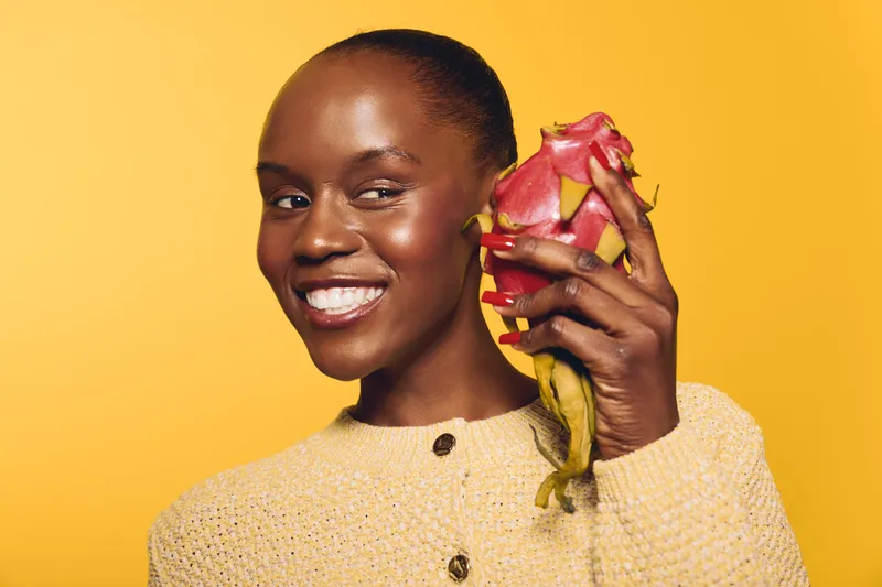 Smiling African woman with dark skin holding dragon fruit near face on yellow background. Concept of healthy lifestyle, fresh fruit, nutrition, and natural beauty.
