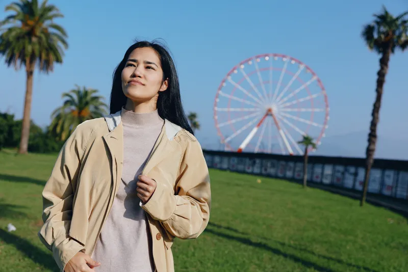 Woman strolling outdoors in a park beside a ferris wheel, palm trees and blue sky, wearing a beige jacket and light sweater, capturing a relaxed sunny day ambiance