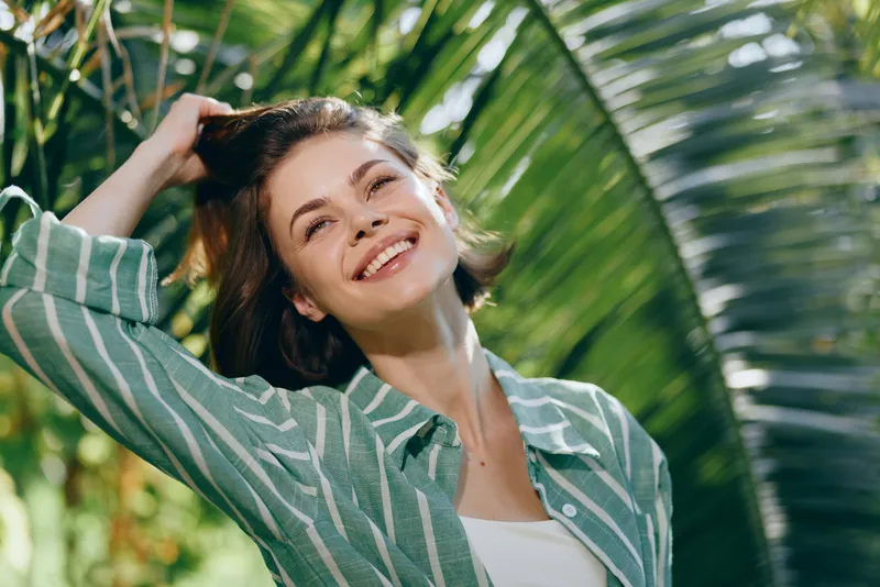 Young woman smiling outdoors, wearing a striped green shirt, captured amid bright sunlight and lush tropical foliage for a fresh, natural portrait.