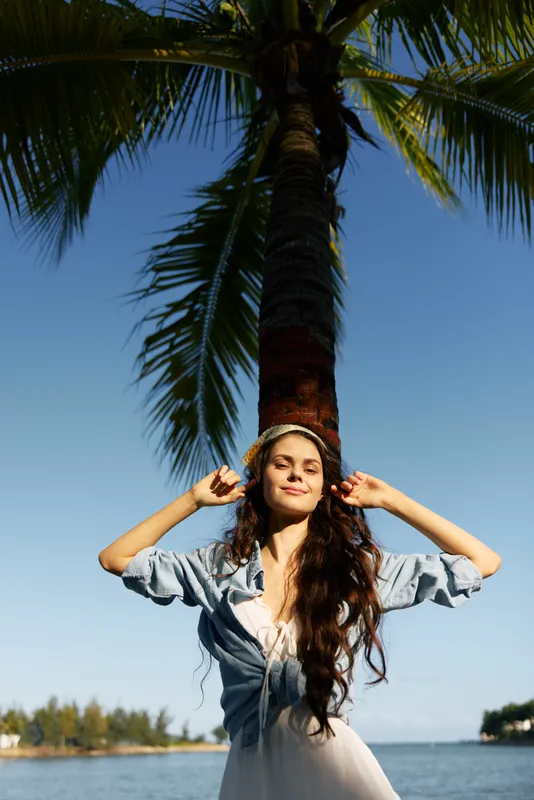 A young woman stands under a tall palm tree by a bright blue sea, arms raised, smiling softly, enjoying sunlight in a relaxed outdoor portrait scene.