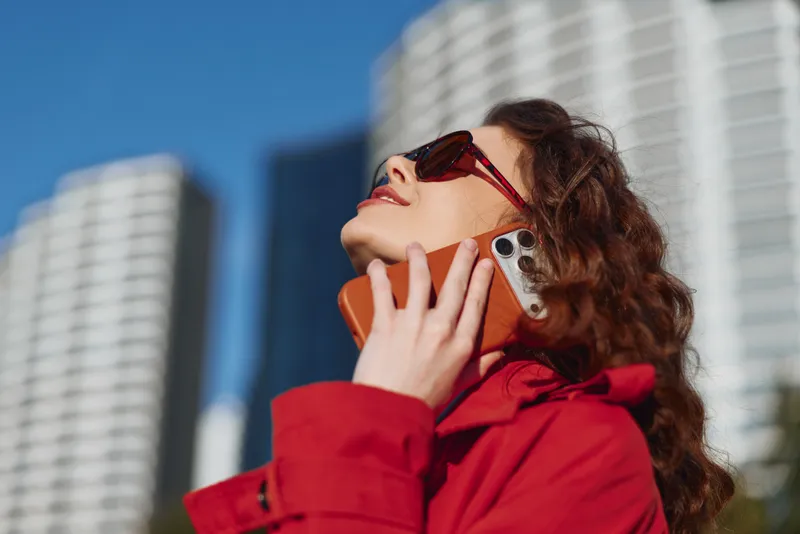 Smiling woman in a red coat talking on a mobile phone outdoors. Urban skyline backdrop with tall buildings. Warm daylight conveys connection, confidence, and city life.