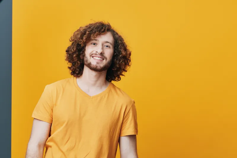 Cheerful young man with curly hair in a bright yellow t shirt against a vibrant orange background, expressing joy and positivity