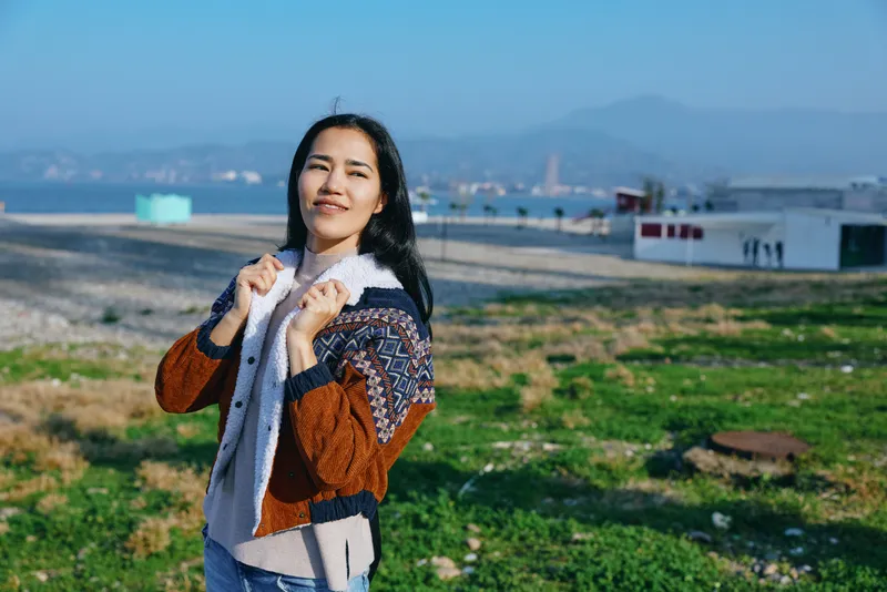 Woman outdoor landscape by sea and mountains, smiling in a warm jacket, standing on a green coastal field with breeze in hair, sunny calm scene highlighting nature and travel mood