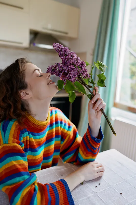 Young woman in a striped multicolor sweater leans over a table indoors to smell a lilac bouquet, capturing a cozy home moment with a warm, intimate vibe, eyes closed.