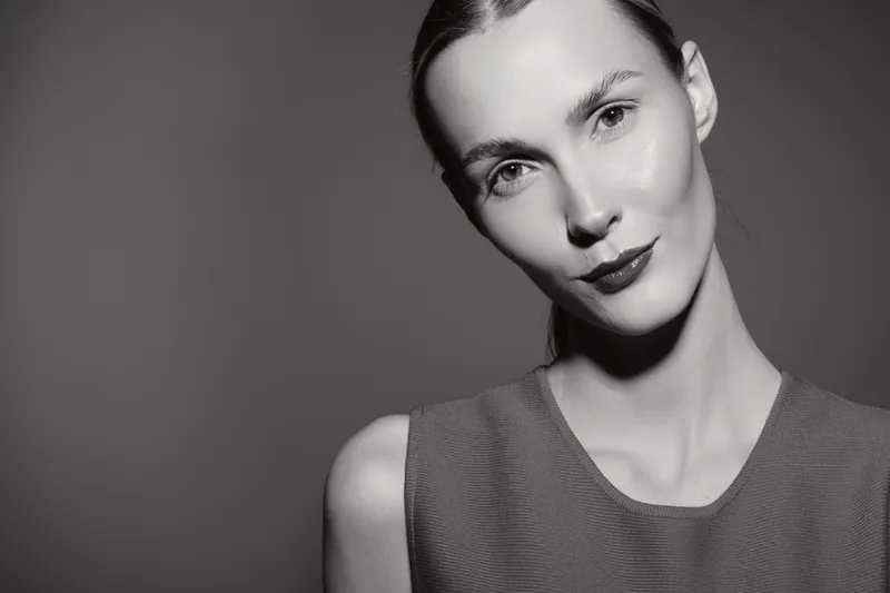 Portrait of a young Caucasian woman with pale skin and slicked back hair wearing sleeveless top on grey background.