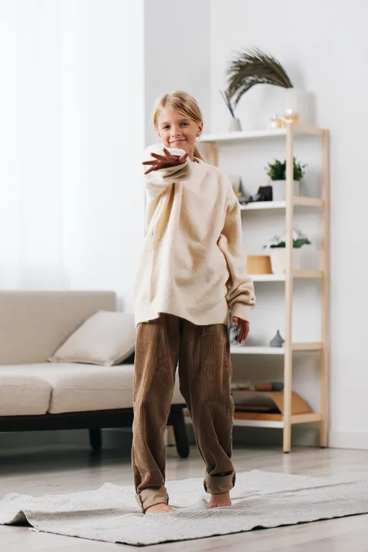 A young girl wearing a soft beige sweater stands in a bright living room, smiling and extending a plate toward the camera in a warm, inviting home scene.