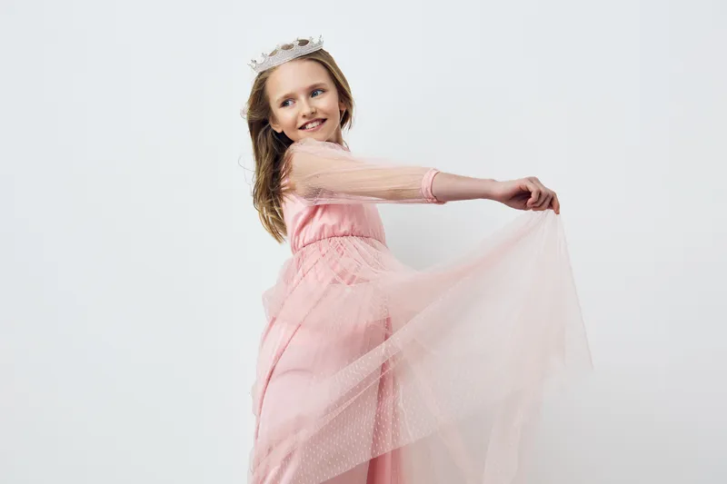Young girl with a crown, wearing a pink dress, joyfully spins in a studio setting, showcasing innocence and happiness in a festive atmosphere