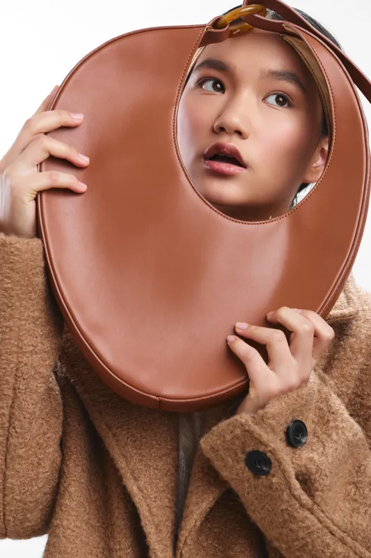 Asian woman wears a cozy coat and peers through a round leather prop, posing in a studio with soft lighting to create a stylish and thoughtful portrait.